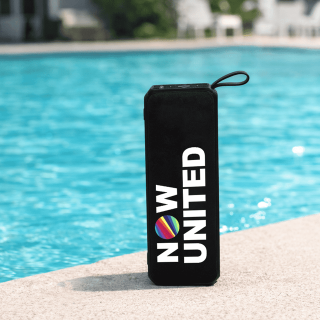 A black portable speaker with a round logo, sitting by a blue pool.