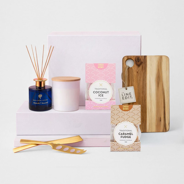 A wooden cheese board, two jars of fudge and coconut ice, a candle, and a diffuser on pink boxes.