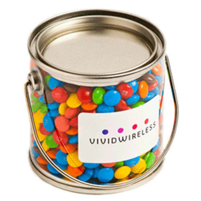 A small silver PVC bucket filled with colourful mini M&Ms in red, blue, green, and yellow. It has a logo on the front.