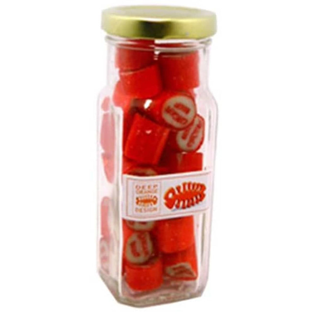 A tall glass jar containing red rock candy, sealed with a gold lid and featuring a branded sticker.
