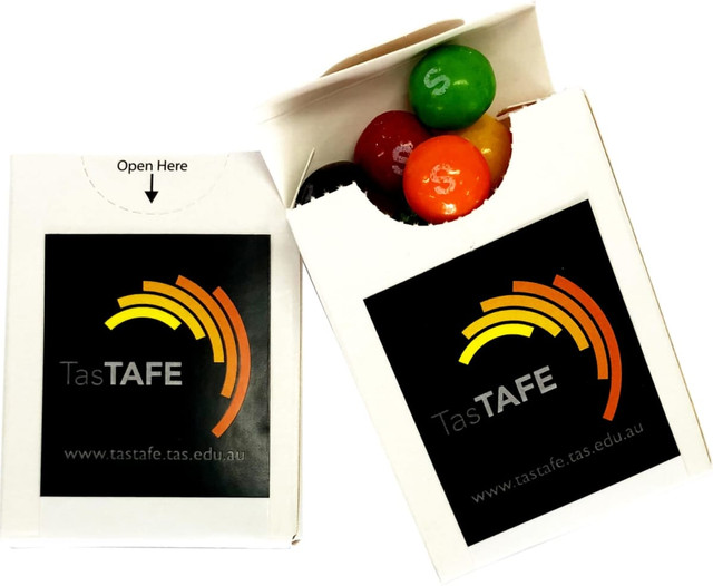 A branded cardboard box containing colourful Skittles, featuring a logo. Box is primarily white with a darker front.
