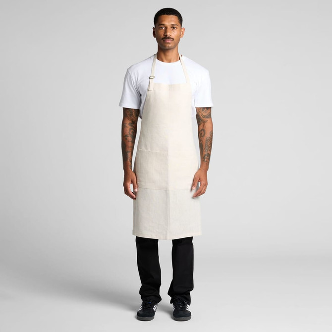 A light beige linen apron worn by a person standing against a neutral background, featuring adjustable straps and a logo.