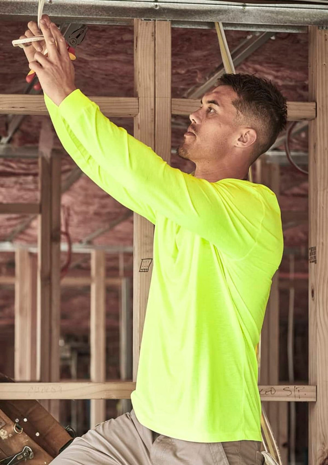 A man wearing a high visibility long sleeve tee in fluorescent yellow, working on electrical wiring in a construction setting.