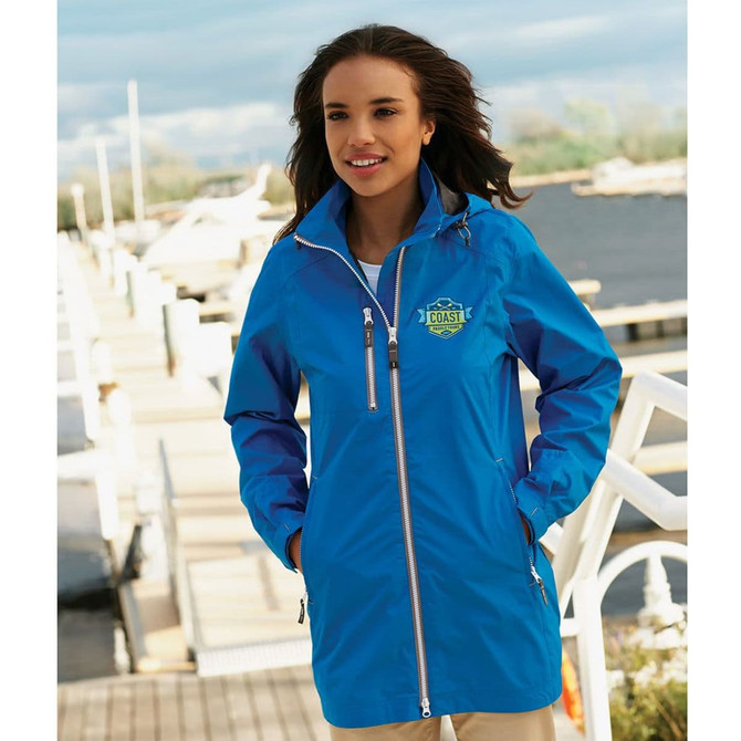 A blue women's rain jacket with a logo, featuring a zip and a hood, set against a scenic waterfront background.