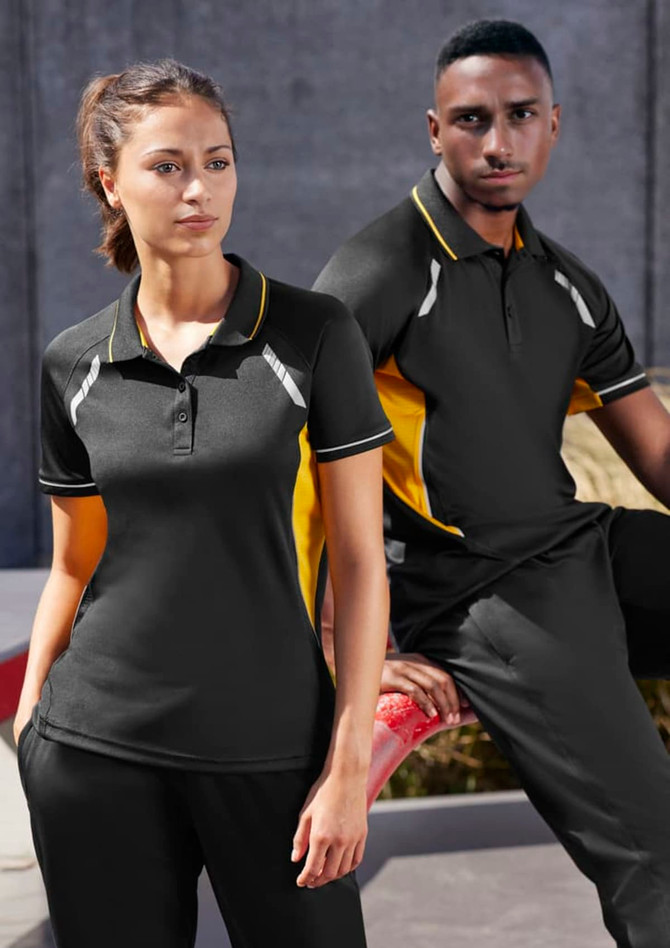 A black and yellow Women's Renegade Short Sleeve Polo, featuring a logo, displayed on a model against an urban backdrop.