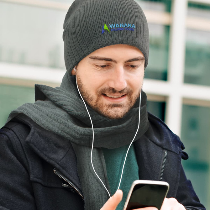 A scarf and beanie set in dark gray featuring a logo on the beanie, worn by a man who is looking at his phone.