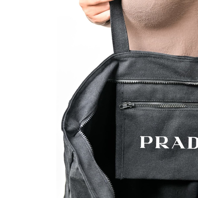 A black tote bag with an interior pocket and a visible logo. The bag's straps are being held by a hand.