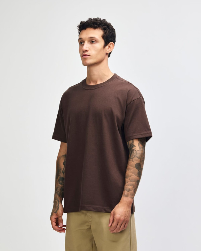 A brown heavyweight t-shirt is held by a model wearing green cargo pants. The shirt has a logo on it.
