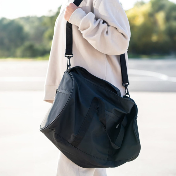 A large black duffle bag with a shoulder strap, carried by a person in a casual outfit against a natural background.