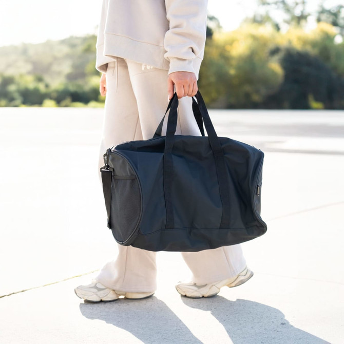 A person carrying a black duffle bag with dual handles, set against an outdoor background.