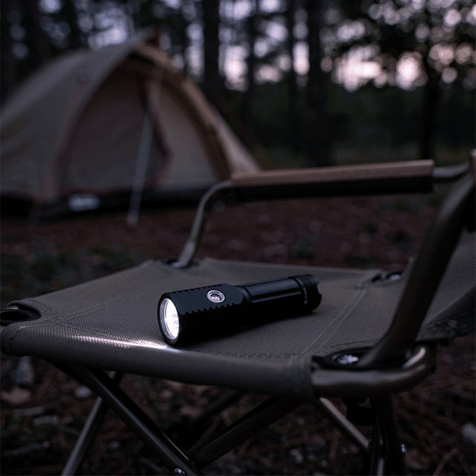 A black LED torch resting on a camping chair, with a tent in the blurred background among trees.