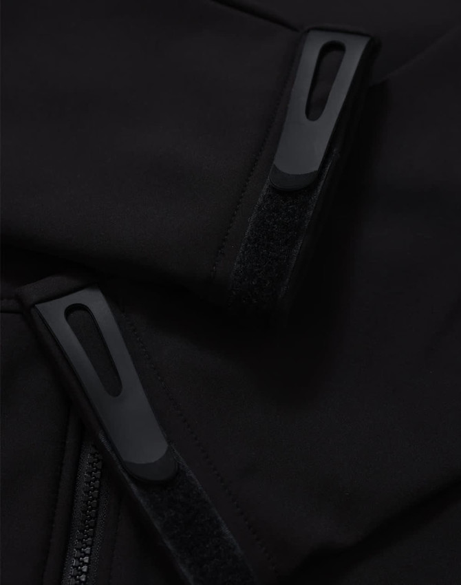 Close-up of a black softshell jacket featuring zippers and velcro details. The jacket has a logo on it.
