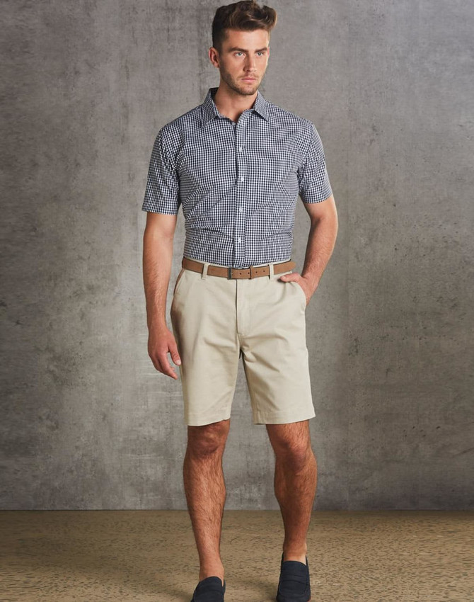Men's chino shorts in beige, paired with a checked short-sleeve shirt and a brown belt. The model stands against a textured grey backdrop.