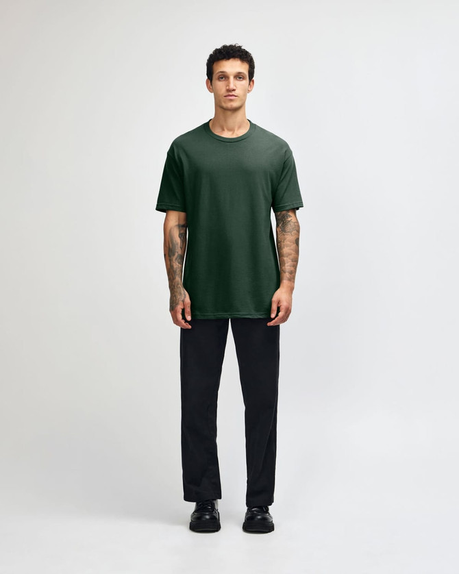 A person wearing a dark green heavyweight cotton unisex T-shirt and light blue cargo pants, standing with hands behind.