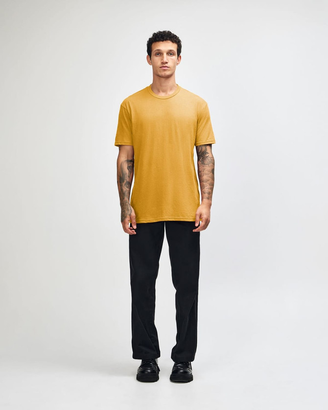 A unisex short sleeve t-shirt in mustard yellow, worn with black cargo pants, featuring a logo.