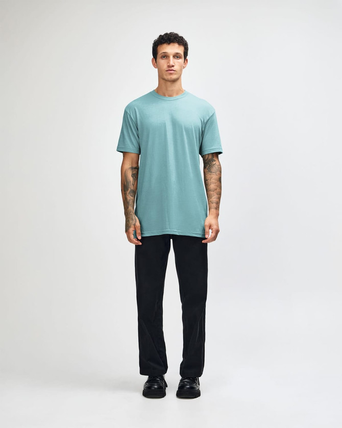 A unisex short sleeve t-shirt in a teal colour, paired with black cargo pants and casual shoes. The model has a relaxed pose.