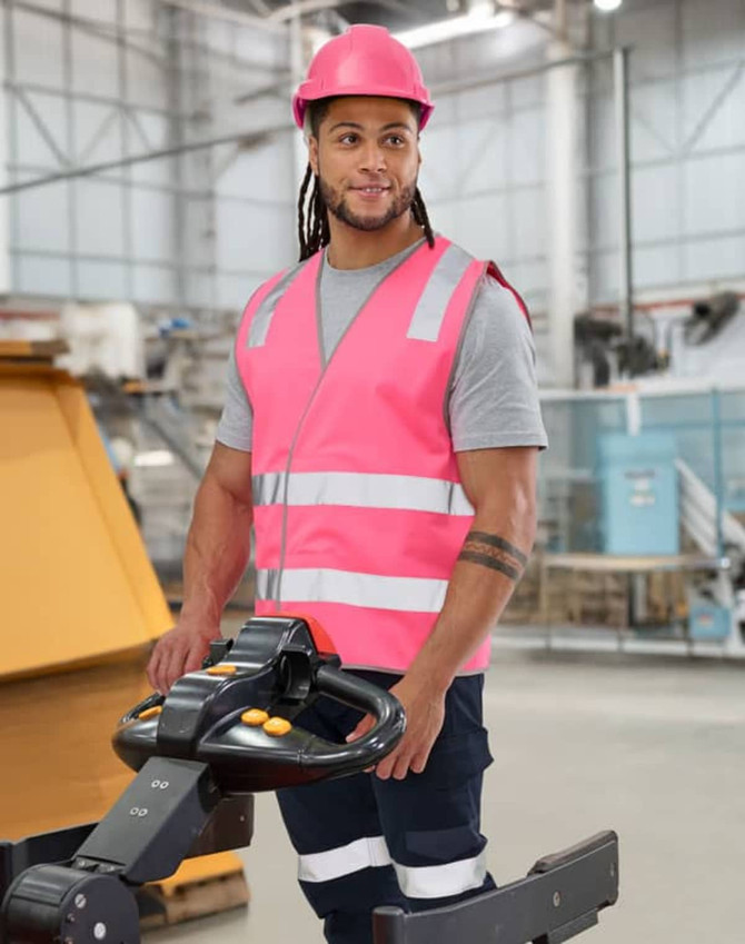 A pink hi-vis safety vest with shoulder reflective tapes, worn by a person in a warehouse setting, featuring a logo.