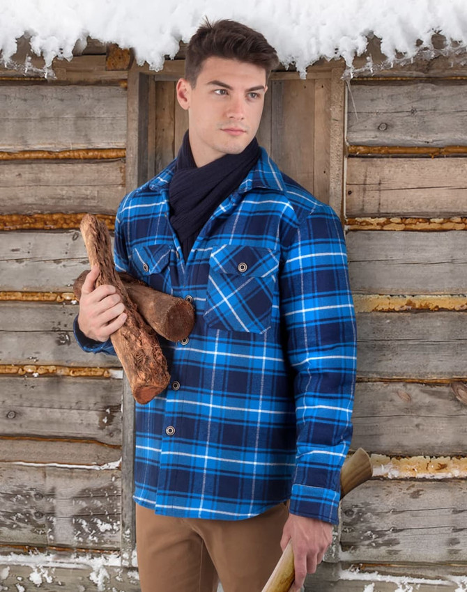 A man stands outdoors in a blue and black quilted flannel shirt, holding firewood near a wooden cabin.