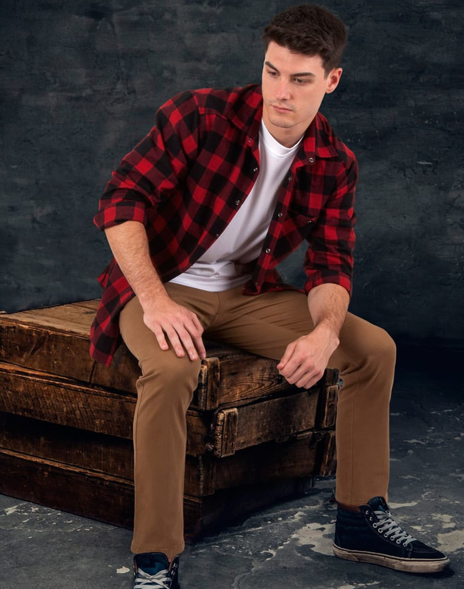 A man sits on a wooden crate wearing a red and black plaid flannel shirt over a white t-shirt with beige pants.