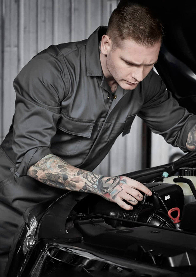 A man wearing a dark grey overall inspects the engine of a car, showcasing detailed tattoos on his arm.