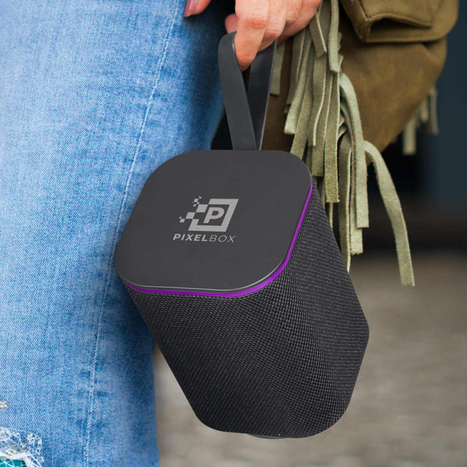 A black Bluetooth speaker with a purple trim and a strap, held by a person wearing denim. It features a logo.