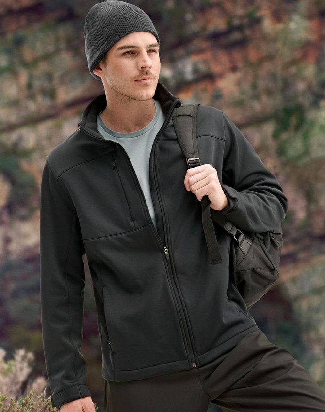 A men's sustainable softshell jacket in black, featuring a zip front and a logo, worn with a beanie and backpack.