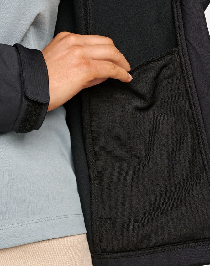 A close-up of a hand reaching into a black pocket on a softshell jacket. The jacket features a contrasting grey layer.