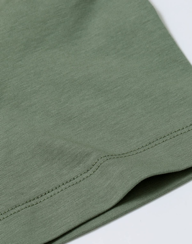 A close-up of a sage green cotton short-sleeve tee shirt featuring a stitched hem and a subtle logo.