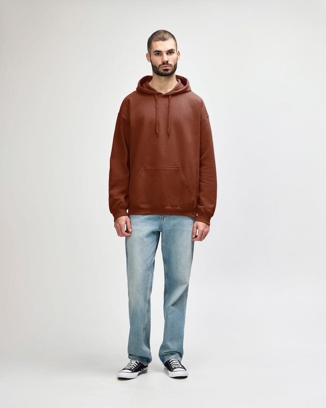 A woman wearing a brown Gildan Softstyle Adult Hooded Sweatshirt and blue jeans, standing against a plain background.