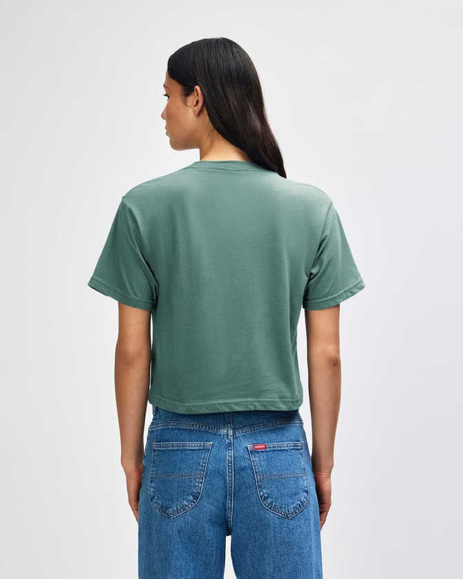 A green women's boxy t-shirt, viewed from the back, paired with light-coloured jeans. It features a branding logo.