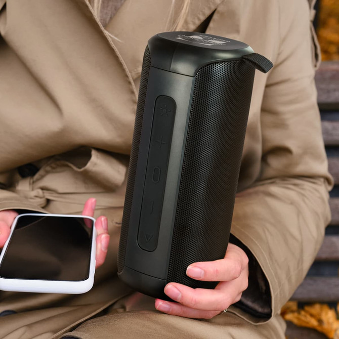 A black Bluetooth speaker with a textured surface, held by a person alongside a smartphone.