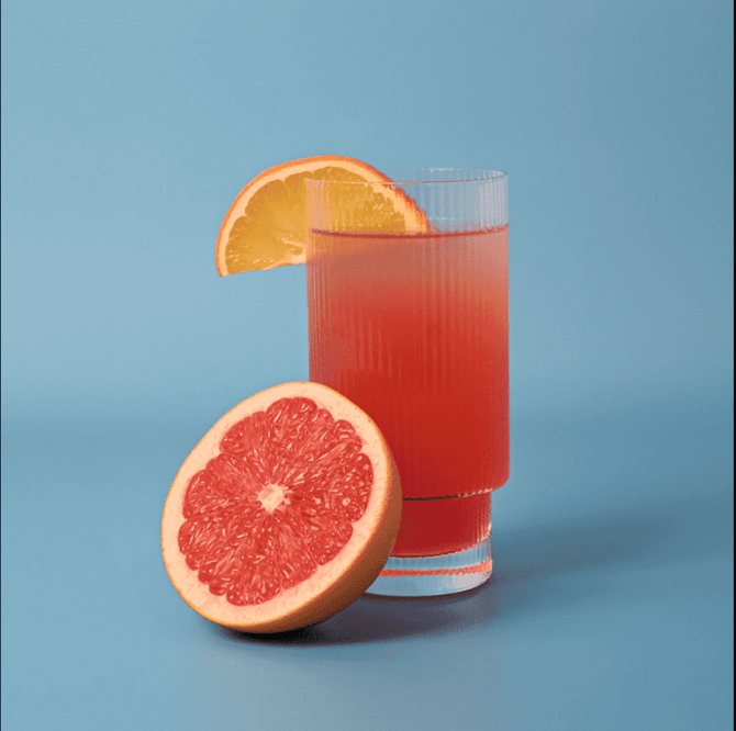A ribbed highball glass filled with a pink drink, garnished with an orange slice, beside a halved grapefruit.
