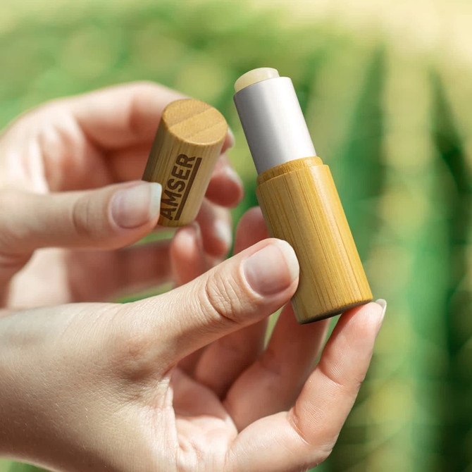 A bamboo lip balm held in hands, featuring a natural wood finish and a white balm core, set against a blurred green background.