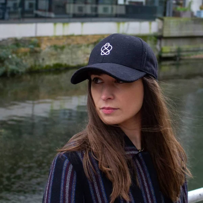 A black heavy acrylic cap featuring a logo, worn by a person beside a body of water.