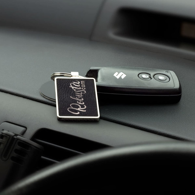 A rectangular key ring in black with a logo, sitting alongside a car key on a dashboard.