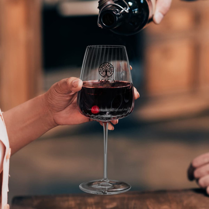 A glass of red wine served in a Luigi Bormioli Optica Bordeaux glass with a logo, held by a person.