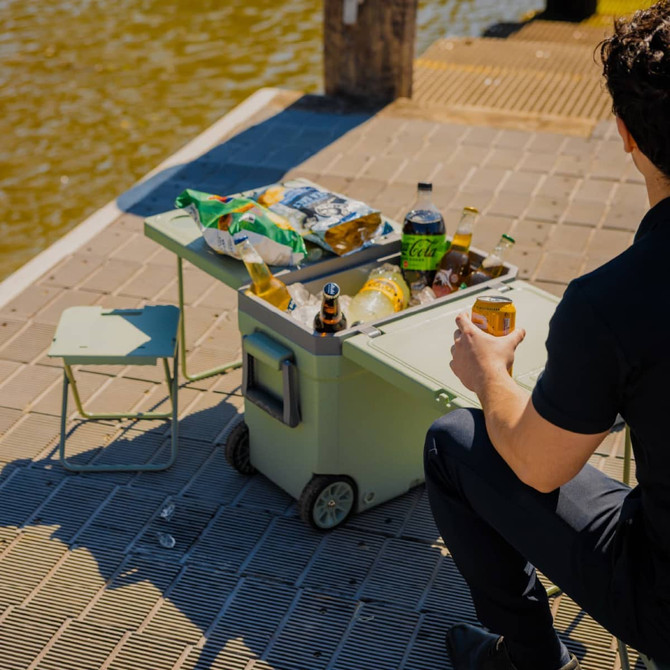 A green cooler box with wheels, featuring a table and a chair, surrounded by snacks and drinks by the water.