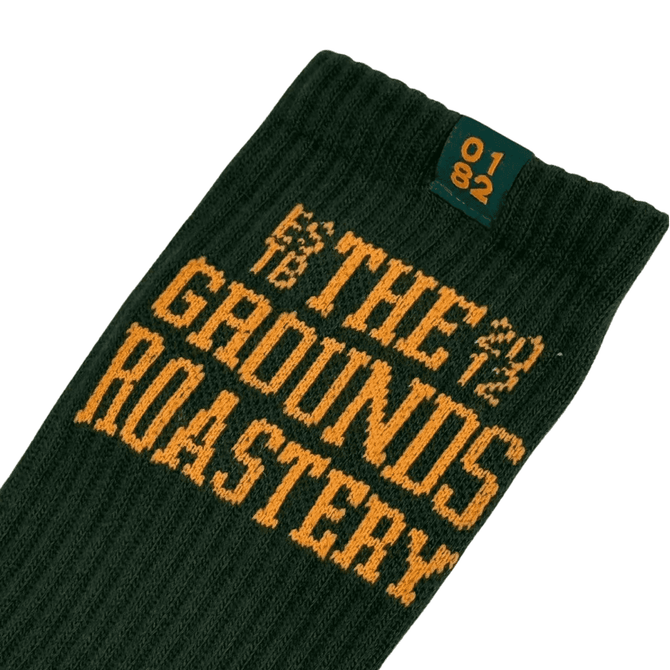 A green sock with yellow text featuring "THE GROUNDS ROASTERY" and a small logo at the top.