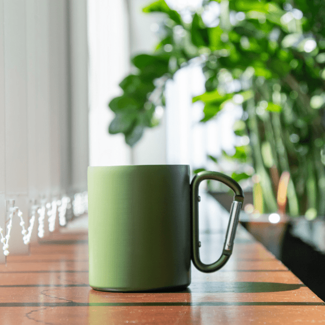 A green thermo mug with a carabiner handle, placed on a wooden surface with plants in the background.