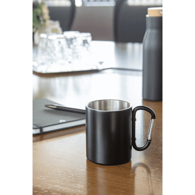 A black thermo mug with a silver interior and a carabiner handle on a wooden table, alongside a bottle and stationery.