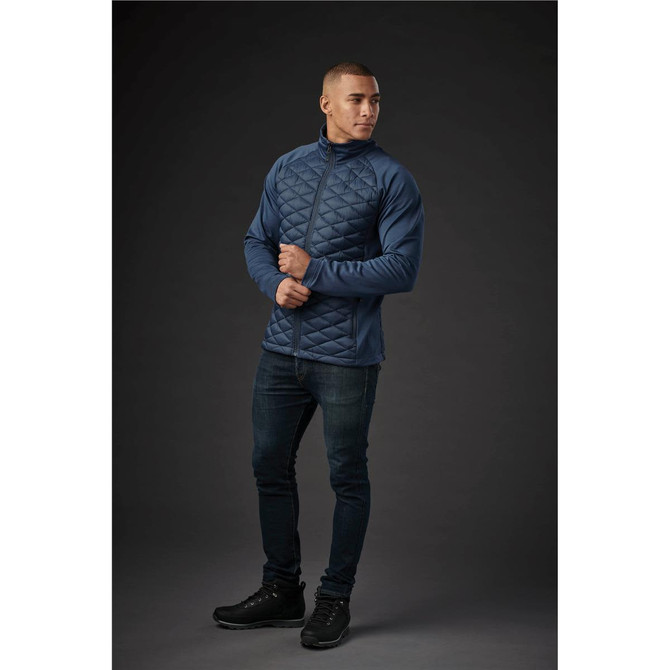 A man wearing a navy blue quilted thermal jacket, paired with dark jeans and black shoes, posing against a dark background.