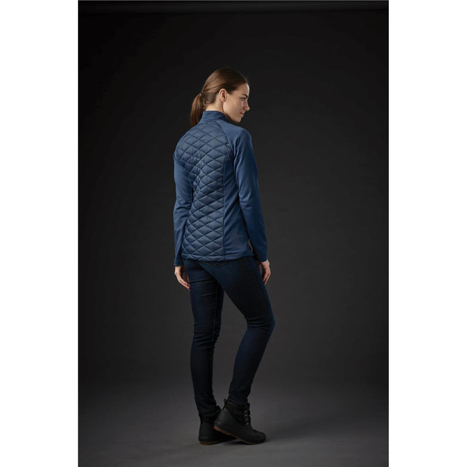 A woman wearing a blue quilted thermal shell jacket, viewed from the back, with dark jeans and footwear.