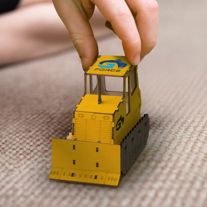 A wooden model of a yellow bulldozer with a logo on top, being manipulated by a hand on a flat surface.