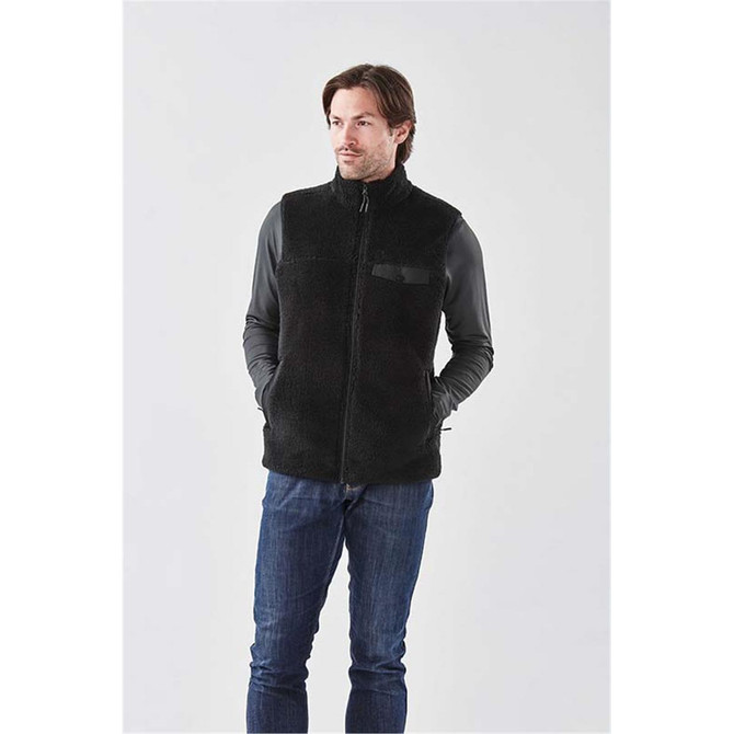 A black men's Sherpa fleece vest features a zip front and a pocket, worn over a long-sleeve top and jeans.