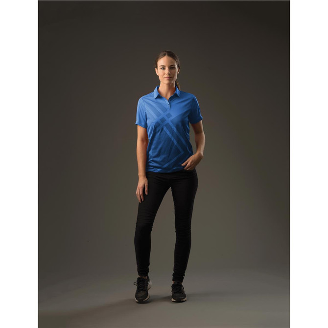 A woman stands wearing a blue Women's Reflex Polo shirt paired with black pants in a studio setting.