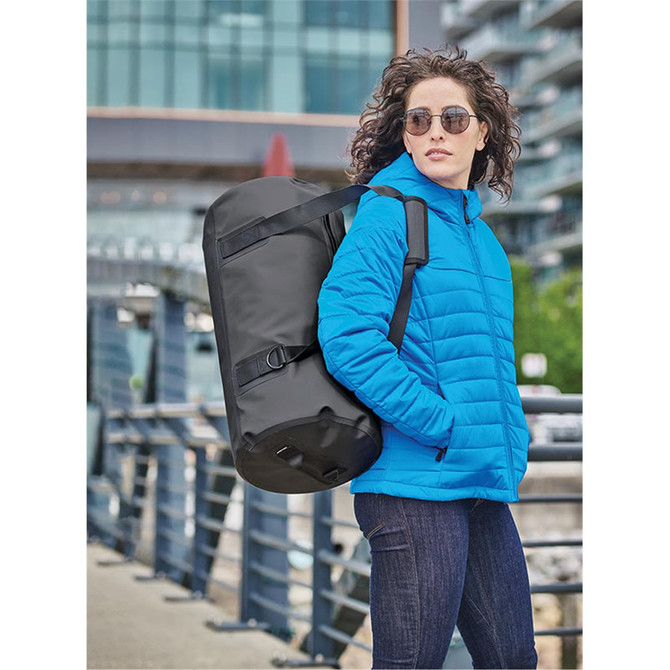 A waterproof duffel bag in black, being carried by a person wearing a blue jacket, with sunglasses on.
