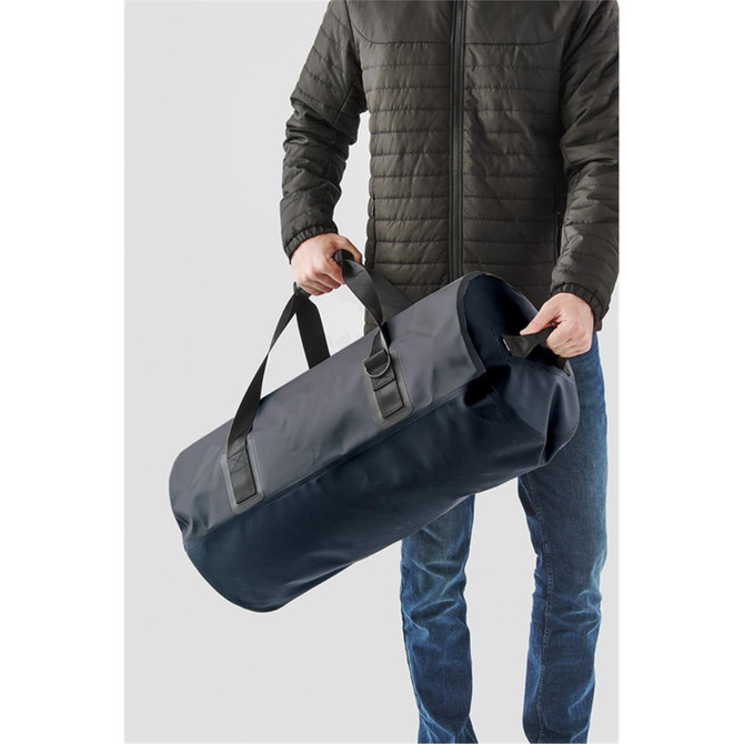 A black waterproof duffel bag being held by a person in a jacket, featuring sturdy handles.