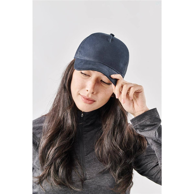 A navy trucker cap rests on the head of a woman with long black hair against a neutral background. The cap features a logo.