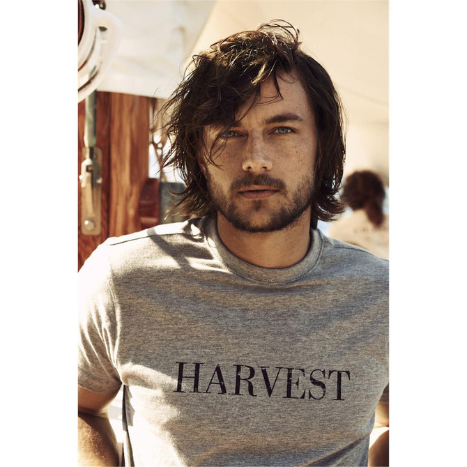 A man wearing a grey crew neck T-shirt featuring a logo. He has long, tousled hair and a beard.