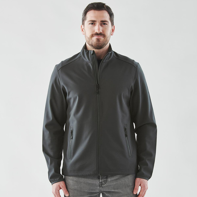 A man wearing a dark grey softshell jacket with a zip front and side pockets. The jacket has a logo.